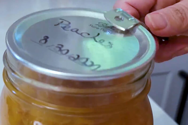 close up of a hand with a can opener opening a mason jar of canned peaches