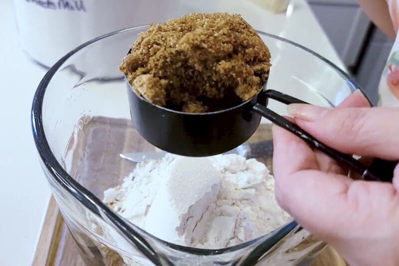 glass bowl with flour and hand holding a measuring cup of brown sugar