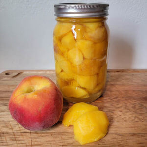 fresh peach and peeled peach used for canning peaches with a jar of canned peaches sitting on a wooden cutting board