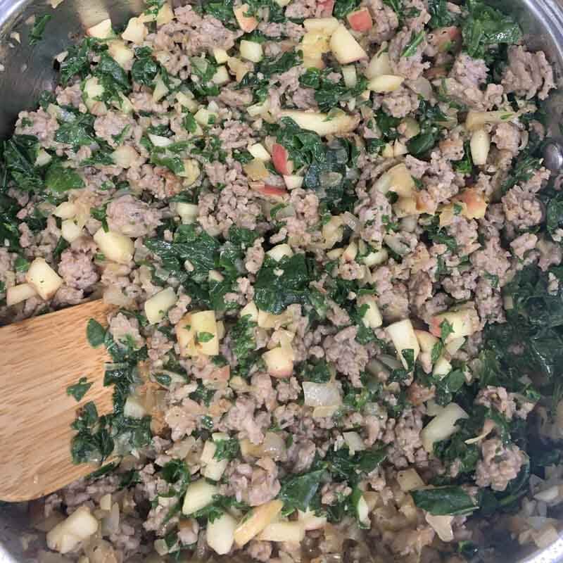 Close up of sausage mixture with apples, onion, and spinach cooking in a pan with a wooden spoon.