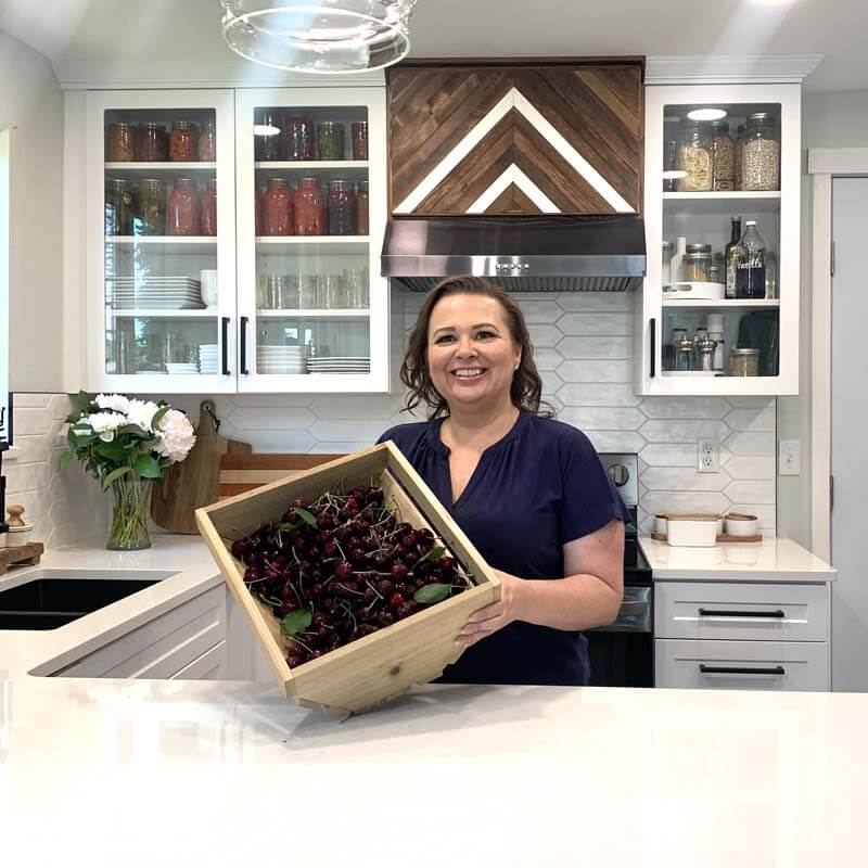 Amy Cross in her kitchen holding a wooden crate full of fresh cherries.
