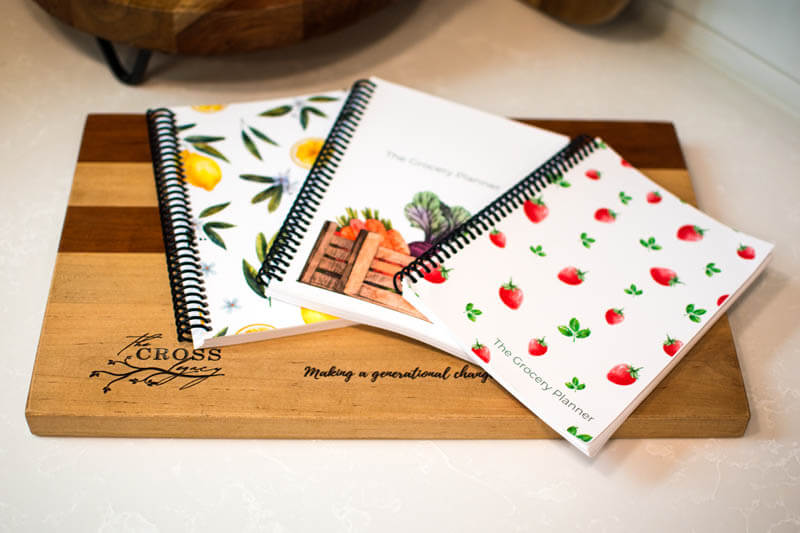 All three The Cross Legacy Grocery Guide and Meal Planners laid out on a wooden cutting board.
